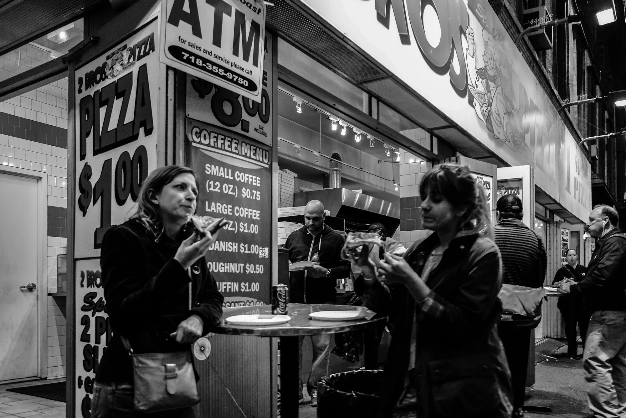 People eating slices outside a 2 Bros. Pizza shop at night